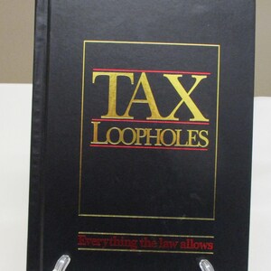 May include: A black book titled "TAX LOOPHOLES" with gold lettering and a red underline. The text "Everything the law allows" is printed in red at the bottom. The book is displayed on a clear stand.