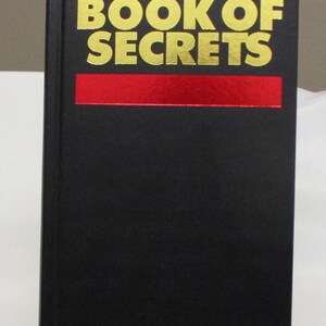 May include: A hardcover book titled "THE BOOK OF SECRETS" with gold lettering on a black cover. The title is centered above a red horizontal stripe. Another red stripe is below the title. The book is displayed on a clear stand.