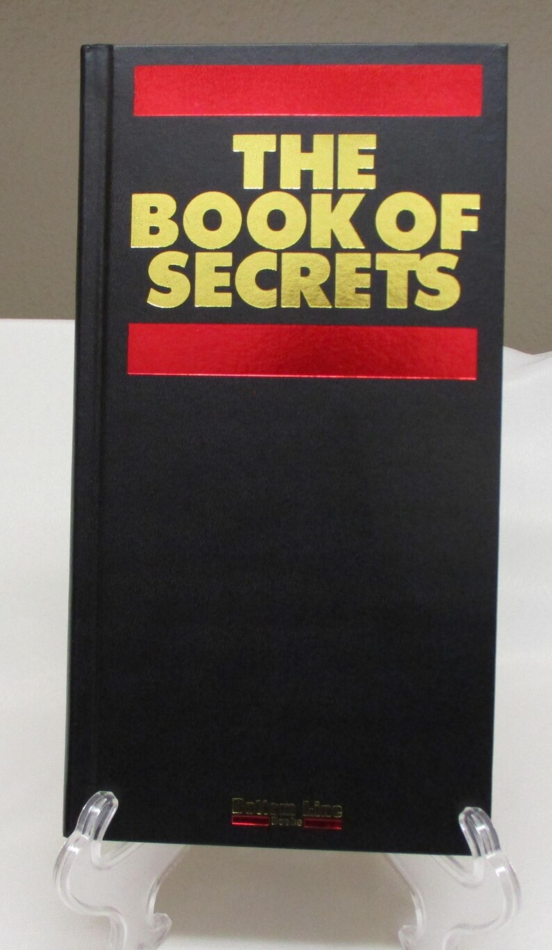 May include: A hardcover book titled "THE BOOK OF SECRETS" with gold lettering on a black cover. The title is centered above a red horizontal stripe. Another red stripe is below the title. The book is displayed on a clear stand.