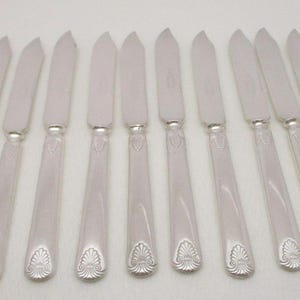 May include: A set of twelve silver butter knives. Each knife has a rounded blade and a handle with a decorative, stylized leaf design at the base. The knives are arranged in a row on a white surface.