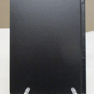 May include: A black book with a textured cover, displayed on a clear plastic stand. The spine is also black, and the book is rectangular in shape. The background is a neutral beige color.