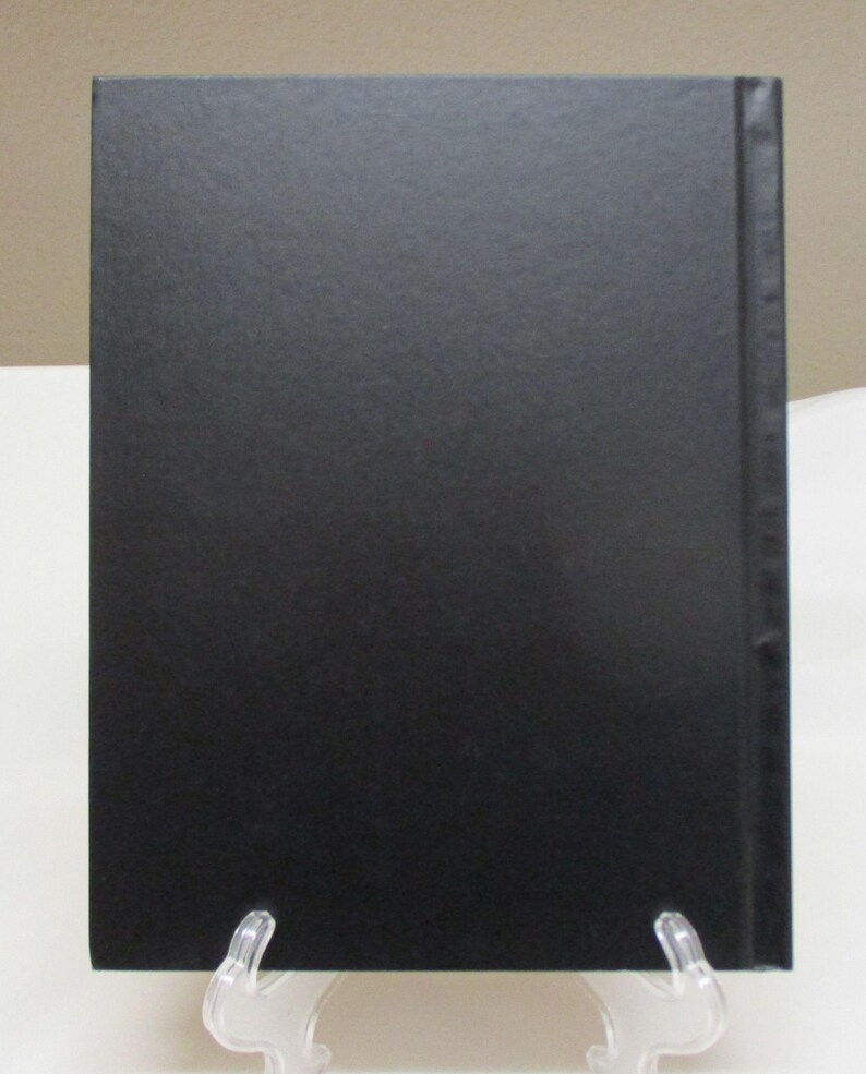 May include: A black book with a textured cover, displayed on a clear plastic stand. The spine is also black, and the book is rectangular in shape. The background is a neutral beige color.