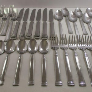 May include: A collection of stainless steel silverware, including forks, knives, and spoons, arranged on a light-colored surface. The silverware has a simple, elegant design with a decorative band near the handle's base. The spoons have an oval bowl.