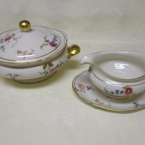 May include: A white porcelain serving dish with a lid and a gravy boat with a matching saucer. Both pieces are decorated with floral patterns in pink, blue, and yellow, and have gold trim. The serving dish has gold handles and a gold knob on the lid.