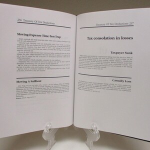 May include: An open book titled "Treasury Of Tax Deductions" with text on the pages. The left page has the title "Moving-Expense Time-Test Trap". The right page has the title "Tax consolation in losses". The book is held open by a clear plastic stand.