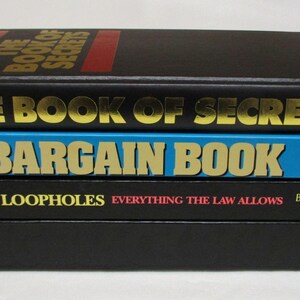 May include: A stack of three books with titles in gold lettering. The top book is black with "THE BOOK OF SECRETS" on the cover. The middle book is blue with "BARGAIN BOOK". The bottom book is black with "TAX LOOPHOLES".
