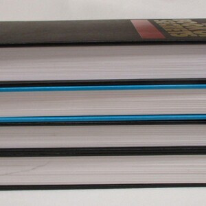 May include: A stack of three hardcover books with white pages and black covers. The top book has a red stripe and gold lettering. The middle book has a blue edge. The books are stacked on top of each other.
