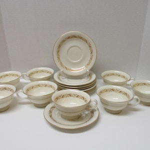 May include: A set of cream-colored teacups and saucers. Each piece features a gold rim and a decorative brown leaf pattern. The set includes eight cups and saucers, and additional saucers.