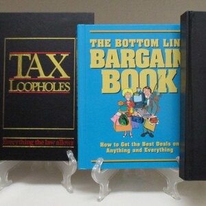 May include: Four books on clear stands. The first book is black with red text that reads "THE BOOK OF SECRETS". The second book is black with gold text that reads "TAX LOOPHOLES". The third book is blue with the text "THE BOTTOM LINE BARGAIN BOOK". The fourth book is black.