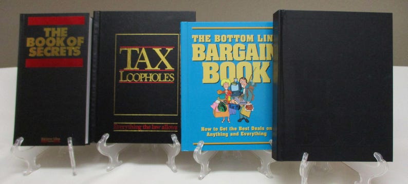 May include: Four books on clear stands. The first book is black with red text that reads "THE BOOK OF SECRETS". The second book is black with gold text that reads "TAX LOOPHOLES". The third book is blue with the text "THE BOTTOM LINE BARGAIN BOOK". The fourth book is black.