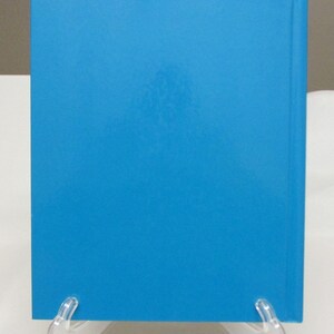 May include: A hardcover book with a bright blue cover. The spine is visible on the right side. The book is displayed on a clear plastic stand against a white background.