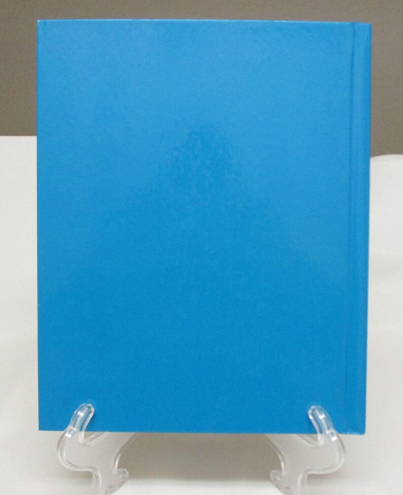 May include: A hardcover book with a bright blue cover. The spine is visible on the right side. The book is displayed on a clear plastic stand against a white background.