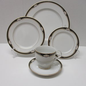 May include: A white porcelain dinnerware set with a black and gold trim, featuring a cup and saucer, and three plates of varying sizes. The set is decorated with a seashell design.