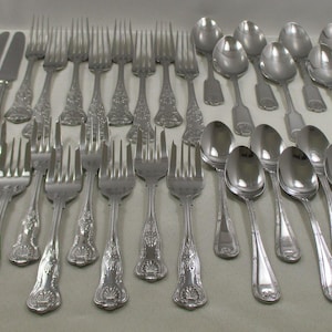 May include: A collection of silver-colored flatware, including knives, forks, and spoons, arranged neatly on a light-colored surface. The silverware has decorative handles and a polished finish, suitable for formal dining.
