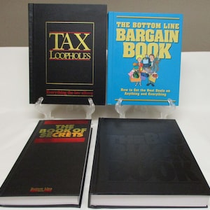 May include: Four books are displayed on clear stands. The books have black and blue covers with titles like "TAX LOOPHOLES" and "THE BOTTOM LINE BARGAIN BOOK." The book "THE BOOK OF SECRETS" is also visible. The books are on a white surface.