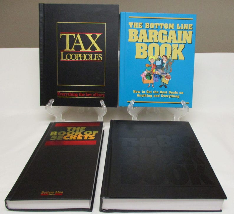 May include: Four books are displayed on clear stands. The books have black and blue covers with titles like "TAX LOOPHOLES" and "THE BOTTOM LINE BARGAIN BOOK." The book "THE BOOK OF SECRETS" is also visible. The books are on a white surface.