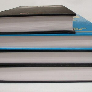 May include: A stack of three hardcover books with white pages and black spines. The top book is black, the middle book is blue, and the bottom book is black. The books are stacked on top of each other.