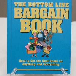 May include: A hardcover book titled "THE BOTTOM LINE BARGAIN BOOK" with a blue cover and yellow lettering. The cover features an illustration of a man and woman carrying shopping bags and other items. The subtitle reads "How to Get the Best Deals on Anything and Everything."