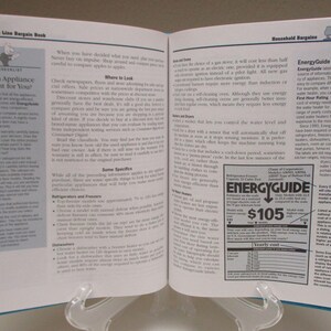 May include: An open book titled "The Bottom Line Bargain Book" with blue covers, displayed on a clear stand. The pages show text and graphics, including an "EnergyGuide" label with a $105 price. The book's focus is on household bargains.