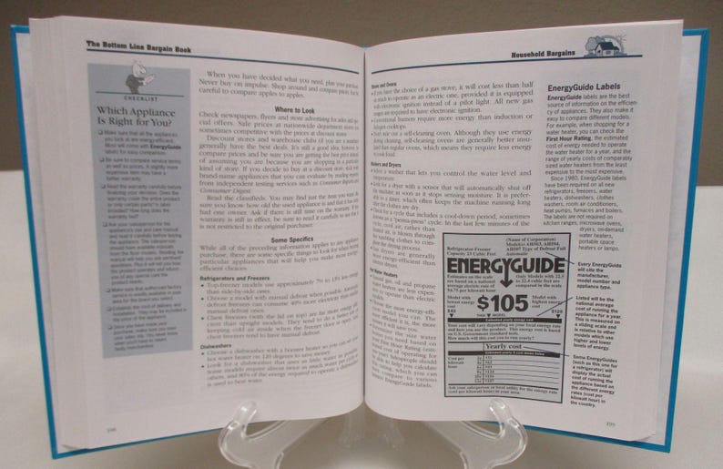 May include: An open book titled "The Bottom Line Bargain Book" with blue covers, displayed on a clear stand. The pages show text and graphics, including an "EnergyGuide" label with a $105 price. The book's focus is on household bargains.