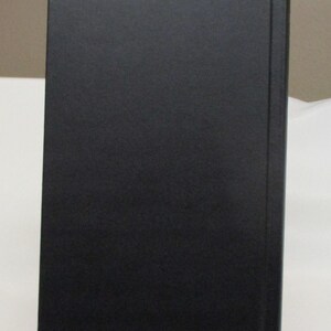 May include: A hardcover book with a solid black cover. The spine is visible on the right side, and the book is propped up on a clear stand. The book's cover has a matte finish.
