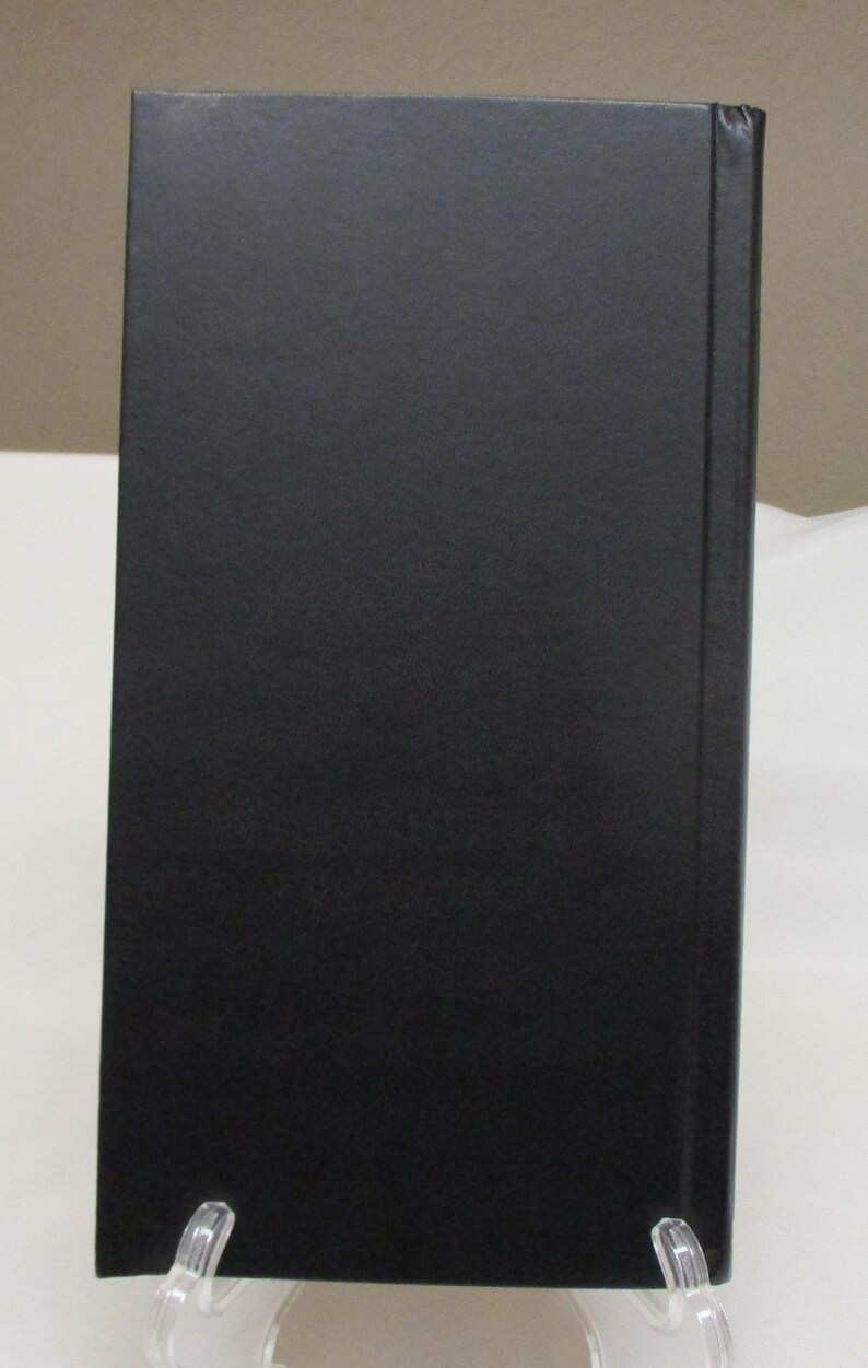 May include: A hardcover book with a solid black cover. The spine is visible on the right side, and the book is propped up on a clear stand. The book's cover has a matte finish.