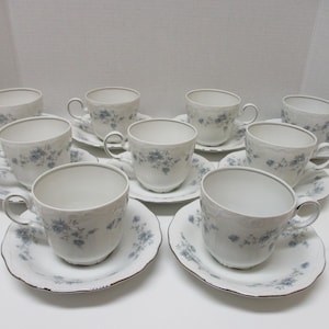 May include: A set of ten white teacups and saucers. Each cup and saucer features a delicate blue floral pattern and a silver trim. The cups have a classic handle design, and the saucers have a scalloped edge.