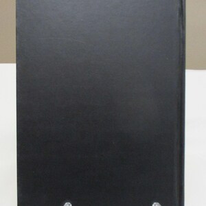 May include: A hardcover book with a black cover. The book is displayed upright on a clear plastic stand. The cover has a matte finish and appears to be made of a textured material.