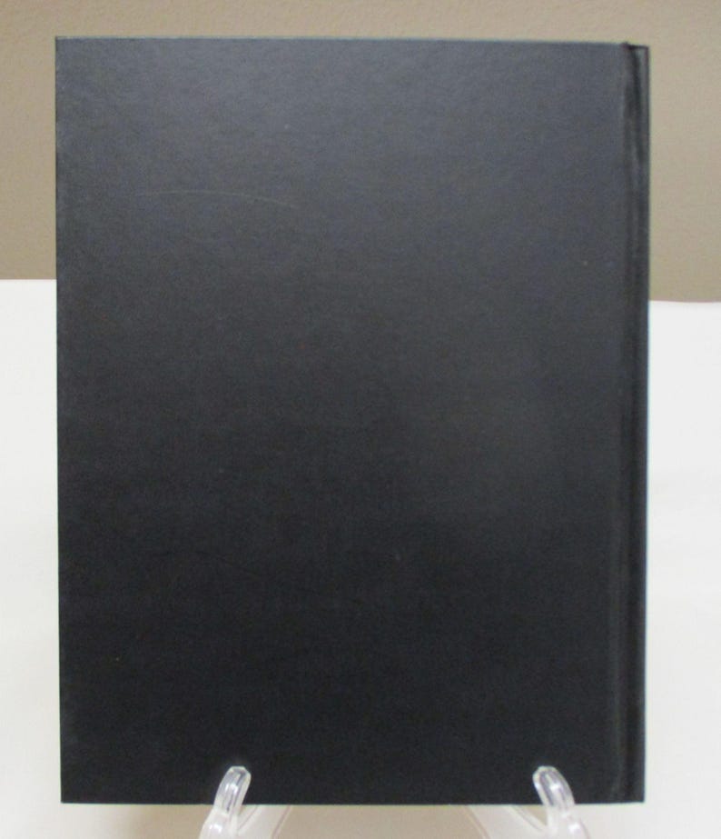 May include: A hardcover book with a black cover. The book is displayed upright on a clear plastic stand. The cover has a matte finish and appears to be made of a textured material.