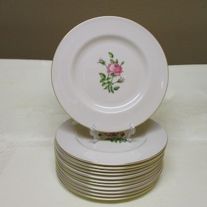 May include: A stack of white ceramic plates with a gold rim. Each plate features a pink rose and green leaves in the center. The plates are stacked on a white surface, with one plate displayed on a clear stand.
