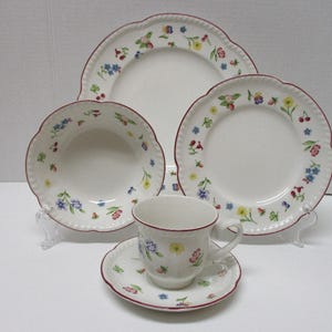 May include: A white floral-patterned dish set, including plates, a bowl, a cup, and a saucer. The set features a scalloped edge with a red trim and colorful flower designs. The set is displayed on clear stands.