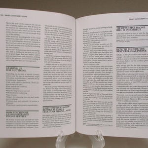 May include: Open book with black cover, pages filled with small text. Headlines include "GEARING UP FOR AUCTIONS" and "WHAT YOU MUST KNOW BEFORE BUYING A CELLULAR PHONE...NOW". The book is propped up on a clear stand.