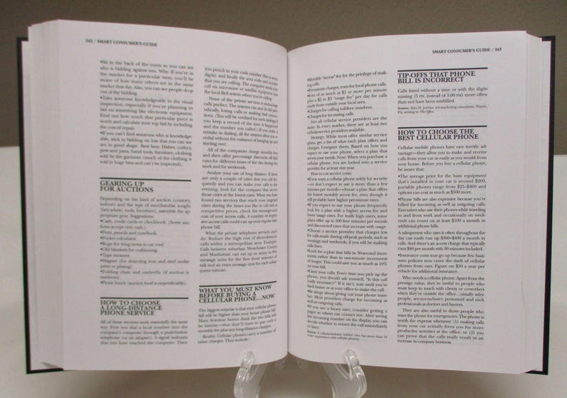 May include: Open book with black cover, pages filled with small text. Headlines include "GEARING UP FOR AUCTIONS" and "WHAT YOU MUST KNOW BEFORE BUYING A CELLULAR PHONE...NOW". The book is propped up on a clear stand.