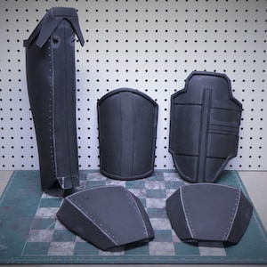 Puede incluir: Un conjunto de piezas de armadura gris oscuro, que incluyen una espinillera, protectores de brazos y protectores de pecho y hombros. Las piezas est&aacute;n dispuestas sobre una superficie a cuadros.