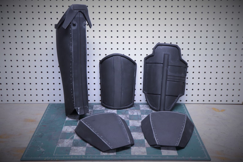 Puede incluir: Un conjunto de piezas de armadura gris oscuro, que incluyen una espinillera, protectores de brazos y protectores de pecho y hombros. Las piezas est&aacute;n dispuestas sobre una superficie a cuadros.