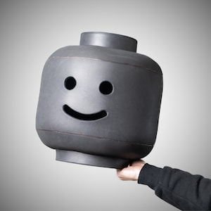 May include: A large, grey Lego-style head with a smiling face. The head features two circular eye holes and a curved mouth. The object is held up by a person's hand, against a grey gradient background.