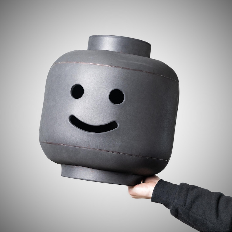 May include: A large, grey Lego-style head with a smiling face. The head features two circular eye holes and a curved mouth. The object is held up by a person's hand, against a grey gradient background.