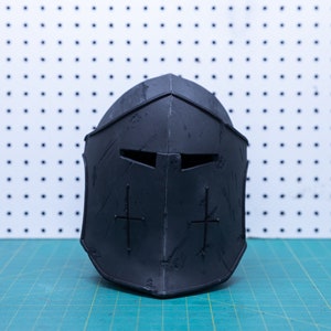 Op de afbeelding: Een zwarte middeleeuwse helm met een verweerde uitstraling. De helm heeft een vizier met twee oogspleten en een kruisontwerp aan elke kant. De helm is gemaakt van donker materiaal en ligt op een groene ondergrond.