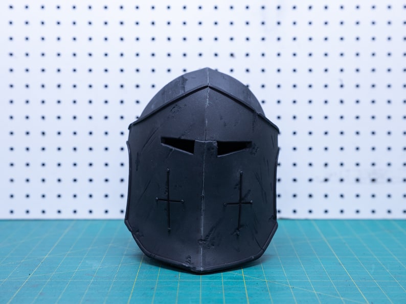 Op de afbeelding: Een zwarte middeleeuwse helm met een verweerde uitstraling. De helm heeft een vizier met twee oogspleten en een kruisontwerp aan elke kant. De helm is gemaakt van donker materiaal en ligt op een groene ondergrond.