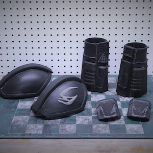 Puede incluir: Un conjunto de piezas de armadura negras de aspecto futurista. El conjunto incluye dos hombreras grandes, dos piezas cil&iacute;ndricas y dos componentes cuadrados m&aacute;s peque&ntilde;os. Las piezas est&aacute;n dispuestas sobre una superficie a cuadros.