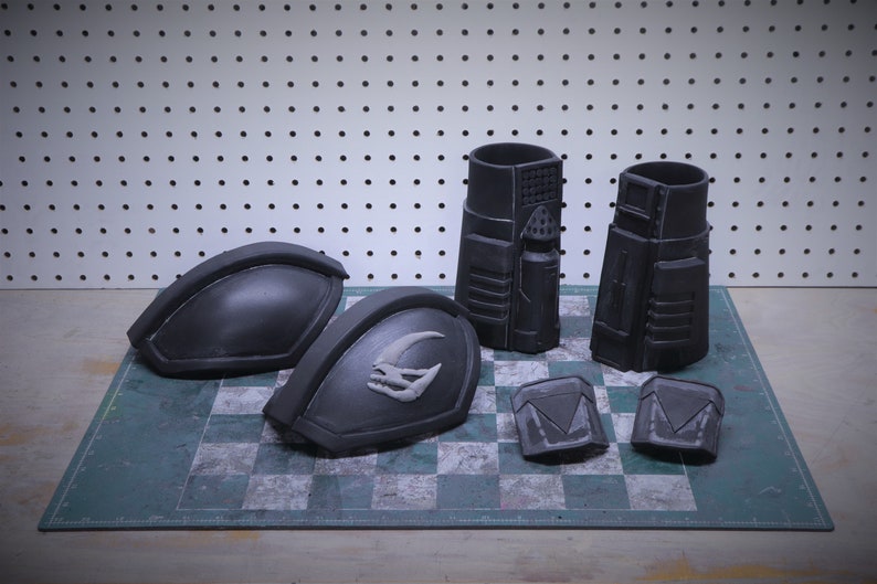 Puede incluir: Un conjunto de piezas de armadura negras de aspecto futurista. El conjunto incluye dos hombreras grandes, dos piezas cil&iacute;ndricas y dos componentes cuadrados m&aacute;s peque&ntilde;os. Las piezas est&aacute;n dispuestas sobre una superficie a cuadros.