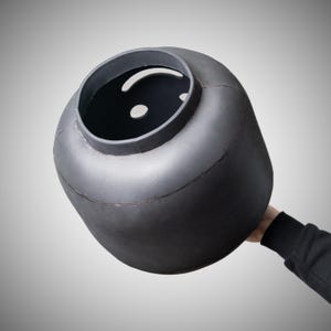 May include: A large, dark grey, cylindrical object with a rounded top and a wide opening. The object has a smooth, matte finish and appears to be made of metal. The object is being held by a person.