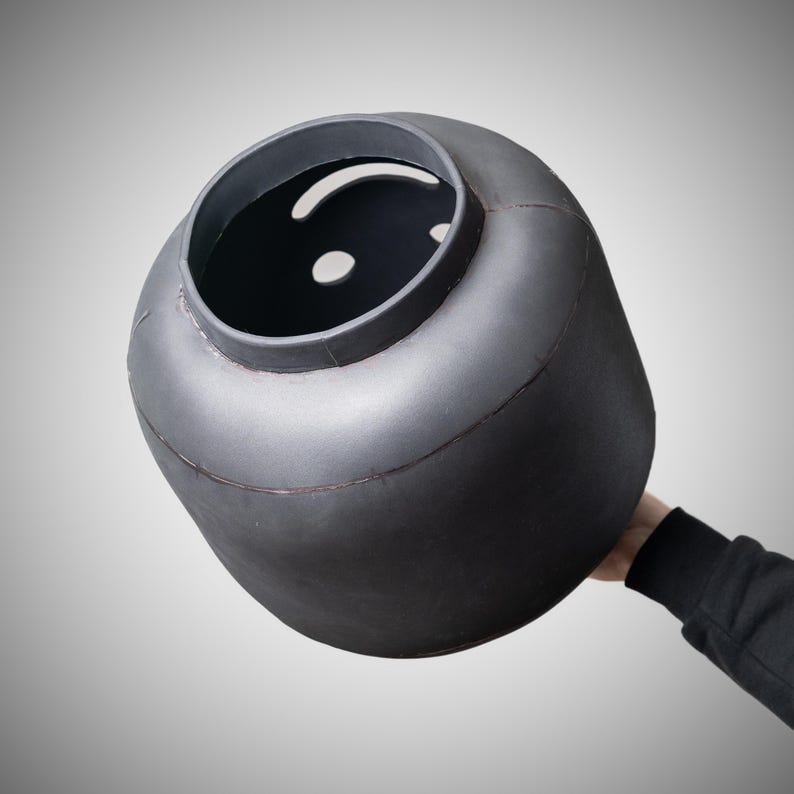 May include: A large, dark grey, cylindrical object with a rounded top and a wide opening. The object has a smooth, matte finish and appears to be made of metal. The object is being held by a person.