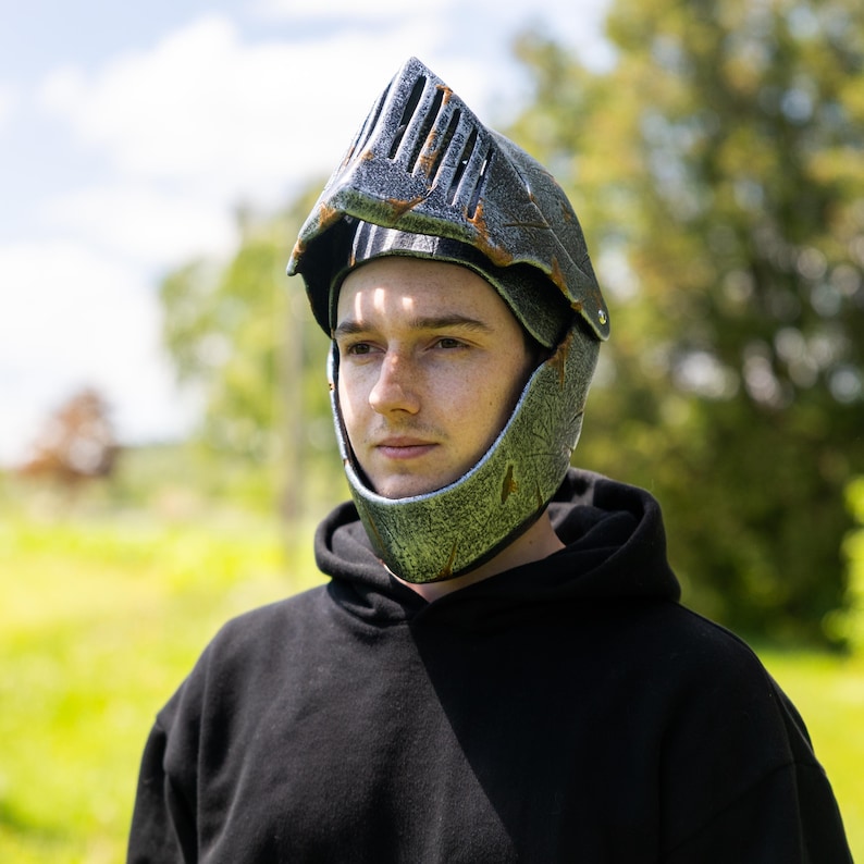 May include: A knight helmet featuring a metallic silver and black weathered finish with rust-colored accents. The helmet includes a pointed visor with vertical ventilation slits and a chin guard, designed to cover the upper face and head area.