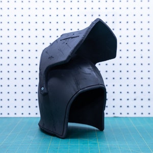 Op de afbeelding: Een zwarte helm met vizier, waarschijnlijk voor cosplay of historische re-enactment. De helm heeft een afgeronde vorm met een prominent vizier en is gemaakt van een mat materiaal. De achtergrond is een wit pegboard en een groene snijmat.