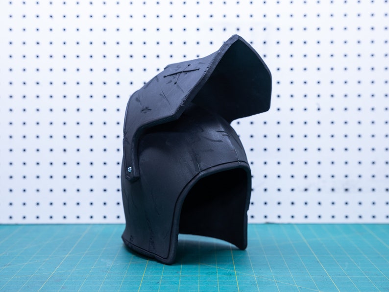 Op de afbeelding: Een zwarte helm met vizier, waarschijnlijk voor cosplay of historische re-enactment. De helm heeft een afgeronde vorm met een prominent vizier en is gemaakt van een mat materiaal. De achtergrond is een wit pegboard en een groene snijmat.