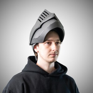 May include: A grey knight helmet constructed from foam material, featuring a pointed visor with horizontal ventilation slits and a curved face guard. The helmet has a structured, angular design with visible seams along the edges and sides.