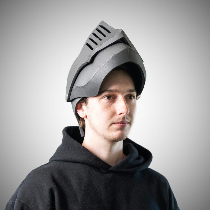 May include: A grey knight helmet constructed from foam material, featuring a pointed visor with horizontal ventilation slits and a curved face guard. The helmet has a structured, angular design with visible seams along the edges and sides.