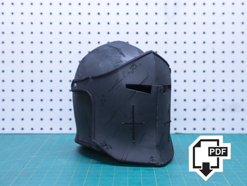 Op de afbeelding: Een zwarte middeleeuwse ridderhelm met een vizier en kruisdetails. De helm is gemaakt van een donker materiaal en heeft een getextureerd oppervlak. De helm ligt op een groene snijmat met een witte pegboard achtergrond.