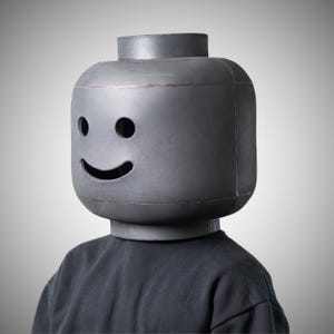 May include: A large, grey Lego-style head mask with a smiling face. The mask has two eye holes and a curved mouth opening. The person is wearing a black long-sleeve top.